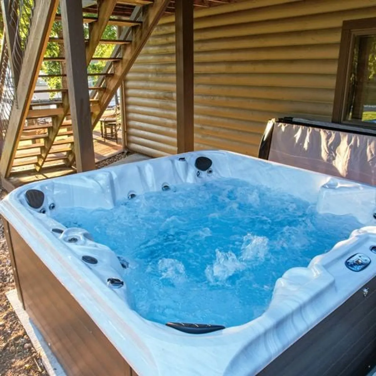 An exterior view featuring a hot tub nestled beside wooden stairs and a log cabin wall, with warm lighting and outdoor seating that suggests a relaxing atmosphere under the open sky.