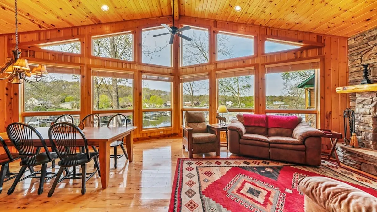 The image showcases a warm and inviting living space with soaring wooden beams, a stunning stone fireplace, and expansive windows that reveal breathtaking lake views. A large dining table and comfortable seating area encourage togetherness and relaxation, while natural light floods the interior, highlighting its rustic charm and modern comforts.