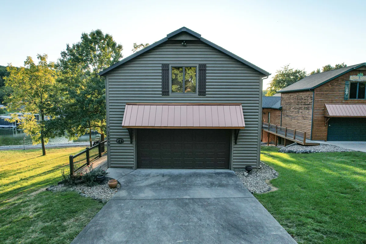 A welcoming exterior view of the house, showcasing the inviting facade, garage door, landscaped lawn, and surrounding trees. The image suggests a homey lakeside environment with plenty of outdoor space for gatherings and relaxation, hinting at resort-style comfort in a private setting.