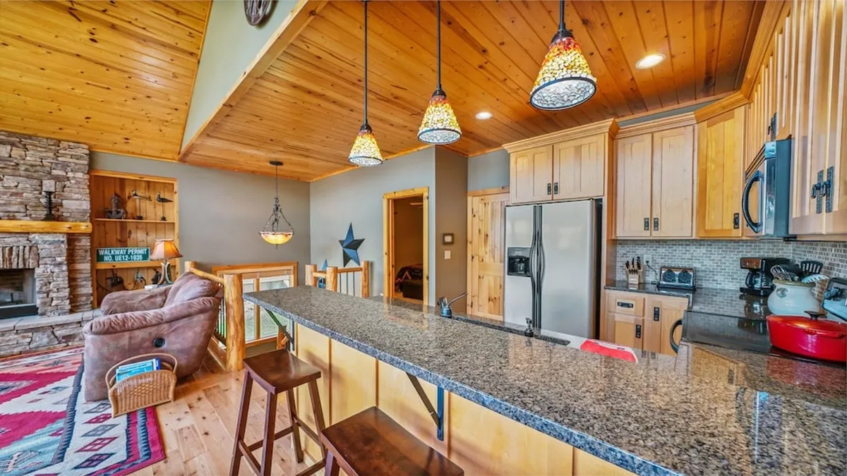 A spacious, warm interior with a stone fireplace, granite countertop, hanging pendant lights, and wooden cabinetry. The scene suggests a place for cooking, entertaining, and family gatherings, highlighting the home’s capacity for comfort and memorable shared moments.