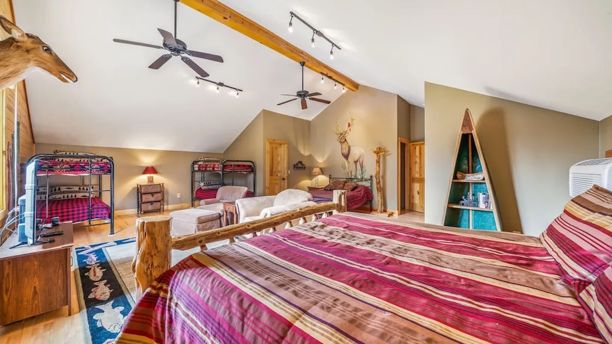 A spacious room featuring a large bed with striped bedding, built-in wooden bunk beds, exposed wooden ceiling beams, a decorative animal head, and a colorful rug. The setup mixes playful and practical, with ample space for kids or families to gather, play, and create memories.
