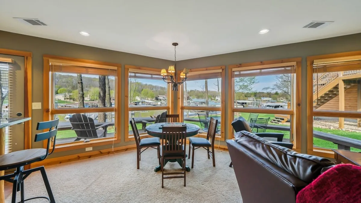 The image shows a round dining table with comfortable seating, set beside large windows that invite in abundant natural light. A chandelier adds a touch of elegance, and the view outside hints at the natural beauty surrounding the lodge. The setup promises a welcoming space for shared meals, games, and conversations.