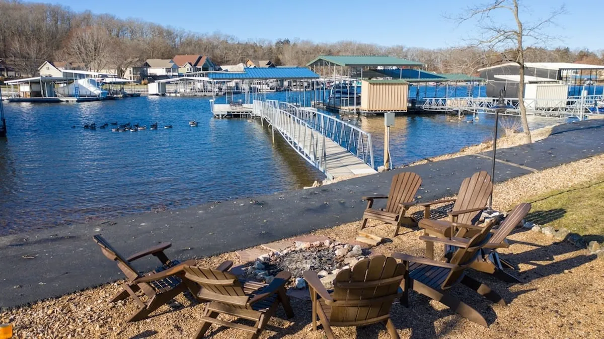 This exterior scene captures a fire pit surrounded by Adirondack chairs on the waterfront, with docks and natural scenery in the background. The setup is ideal for communal activities such as evening campfires, shared meals, and waterside gatherings.