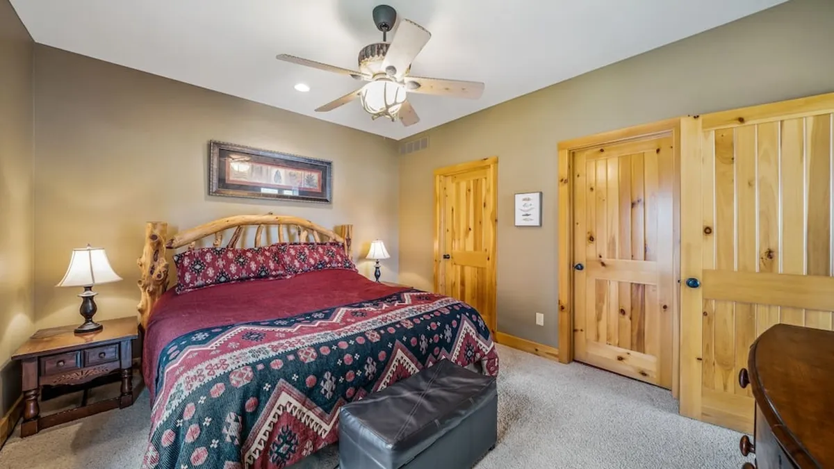 This image features a bed with patterned bedding, wooden furniture, a ceiling fan, and warm light fixtures. The room is filled with soft natural light, creating a calm and restful atmosphere ideal for unwinding after a long day. The overall aesthetic is cozy and welcoming, highlighting comfort and tranquility.
