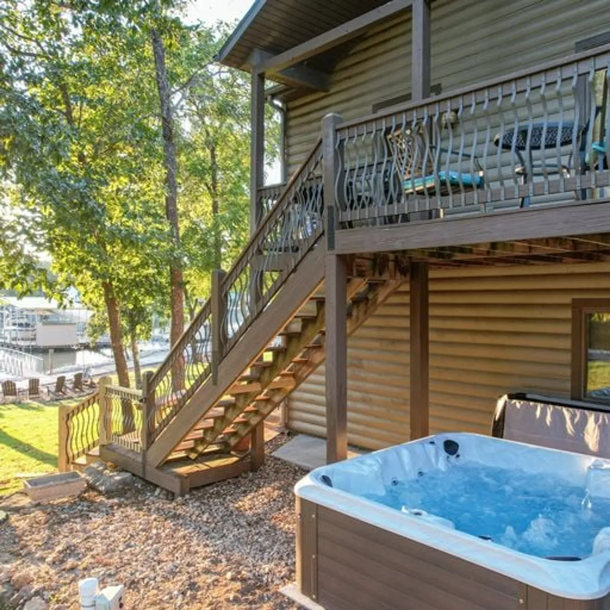 A spacious deck area with a private hot tub set amidst lush greenery. The scene highlights relaxation, with the hot tub positioned to take in the surrounding natural beauty, including glimpses of the lake.