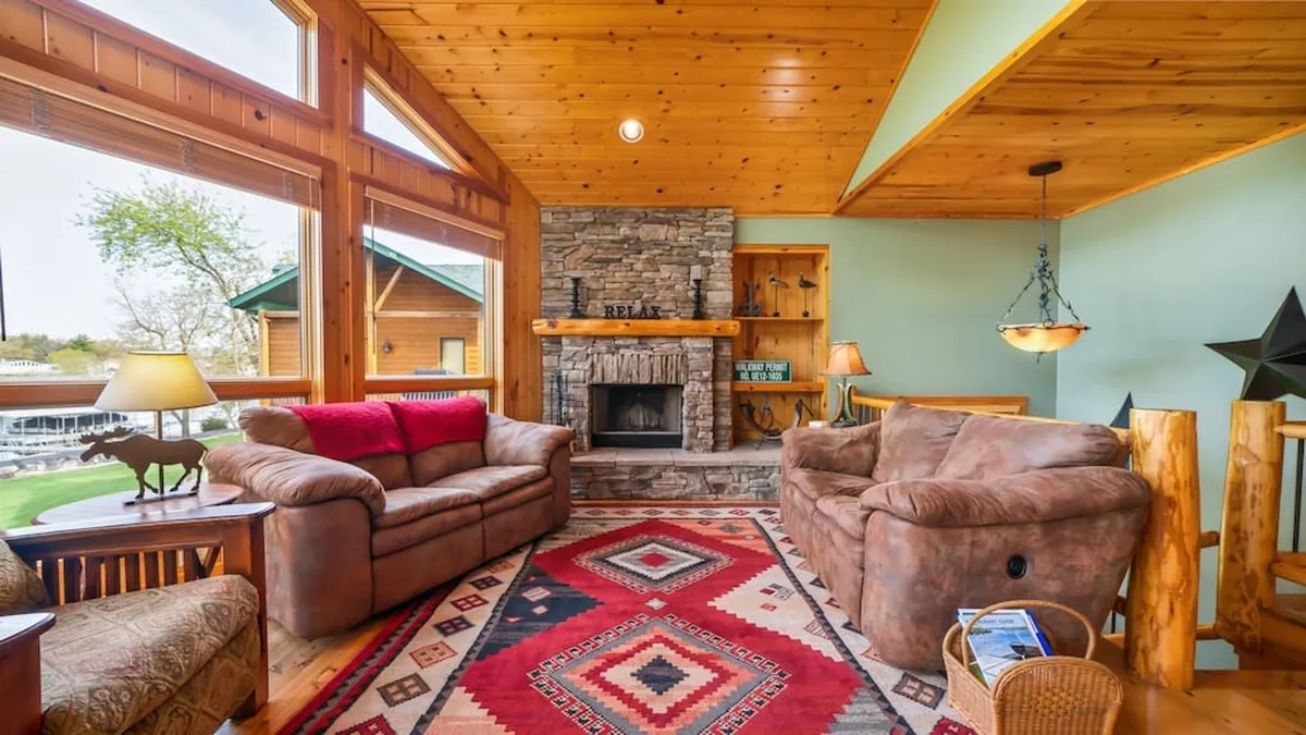 A welcoming living space featuring a cozy fireplace, rug, large windows that let in abundant natural light, and comfortable sofas arranged for relaxation and conversation. The view outside hints at the proximity to nature and the lake.