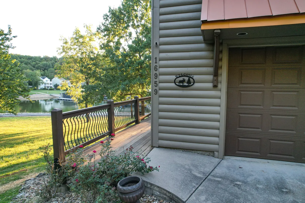 A wide porch with a wooden railing and a cheerful 'Welcome' sign overlooks landscaped gardens and offers a distant view of the water. The setting feels inviting and perfect for gatherings or quiet relaxation.