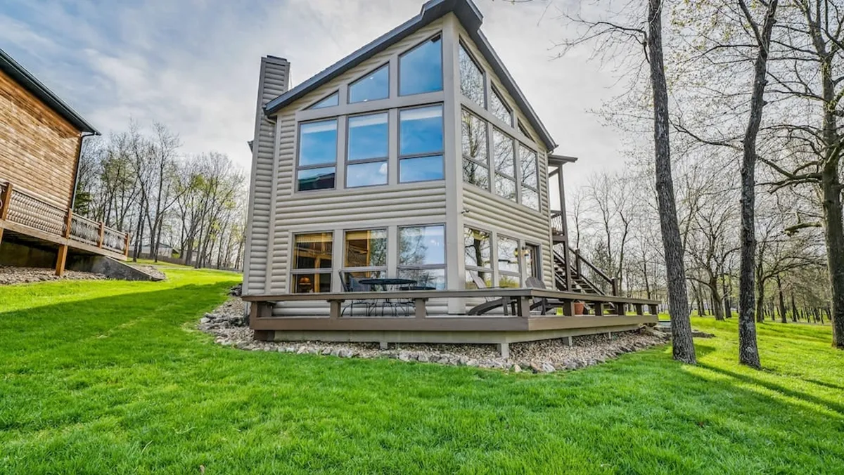 This image features the exterior of Elk Lodge, highlighting its large glass windows, expansive green lawn, and a spacious wooden deck. The scene is peaceful and inviting, suggesting ample space for outdoor relaxation and family gatherings.
