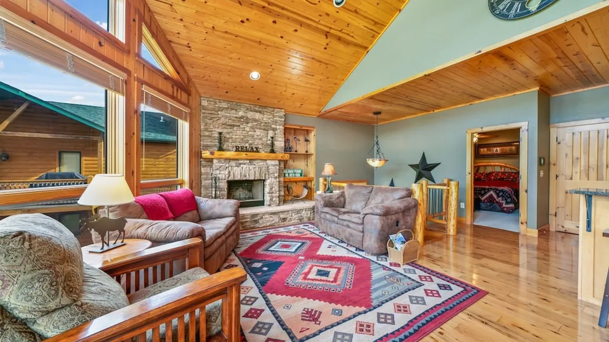 A spacious main floor living room featuring a plush sofa set gathered around a central rug and fireplace. Tall windows offer sweeping lake views, filling the room with natural light. The setting exudes comfort, ideal for relaxing, reading, or socializing with friends and family.